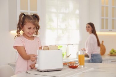 Photo of Cute little girl using toaster at table in kitchen