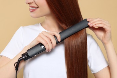Young woman using hair iron on beige background, closeup