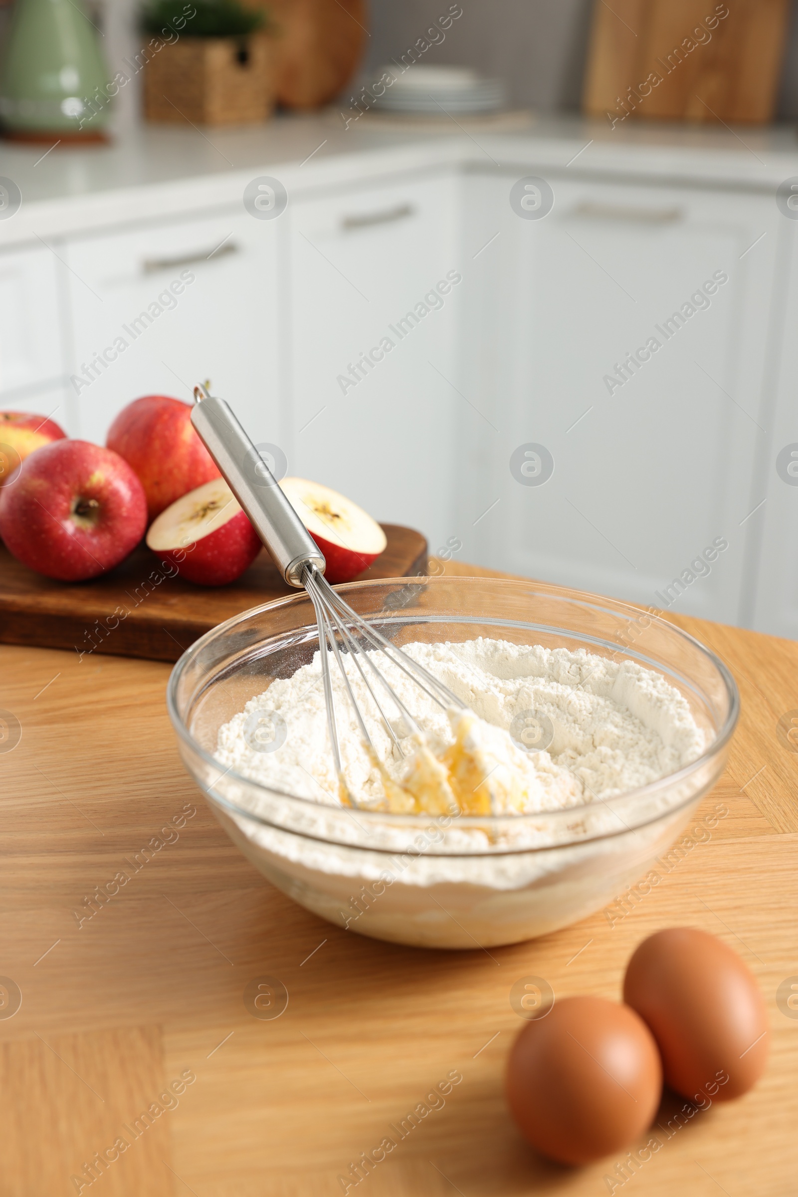 Photo of Cooking process. Metal whisk, bowl, flour, eggs and apple on wooden table in kitchen