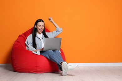 Photo of Cheerful woman with laptop sitting on beanbag chair near orange wall, space for text