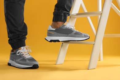 Photo of Man in stylish sneakers standing on white ladder against yellow background, closeup