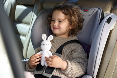 Cute little girl with toy rabbit sitting in child safety seat inside car