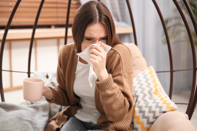 Photo of Sick young woman with cup of hot drink sneezing at home. Influenza virus