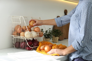 Photo of Woman putting golden onion into storage organizer at countertop in kitchen, closeup