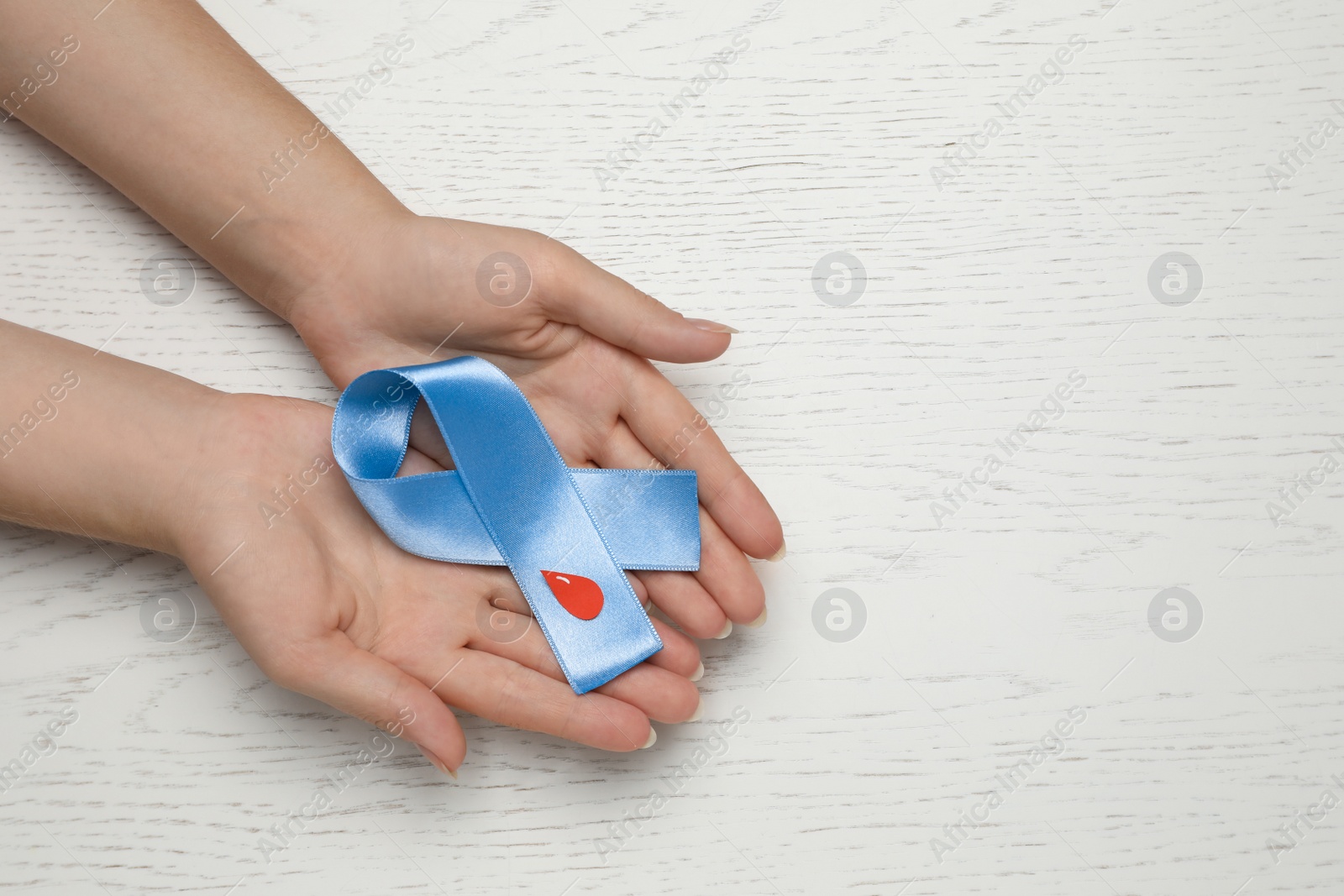 Photo of Woman holding light blue ribbon with paper blood drop at wooden table, top view and space for text. Diabetes awareness