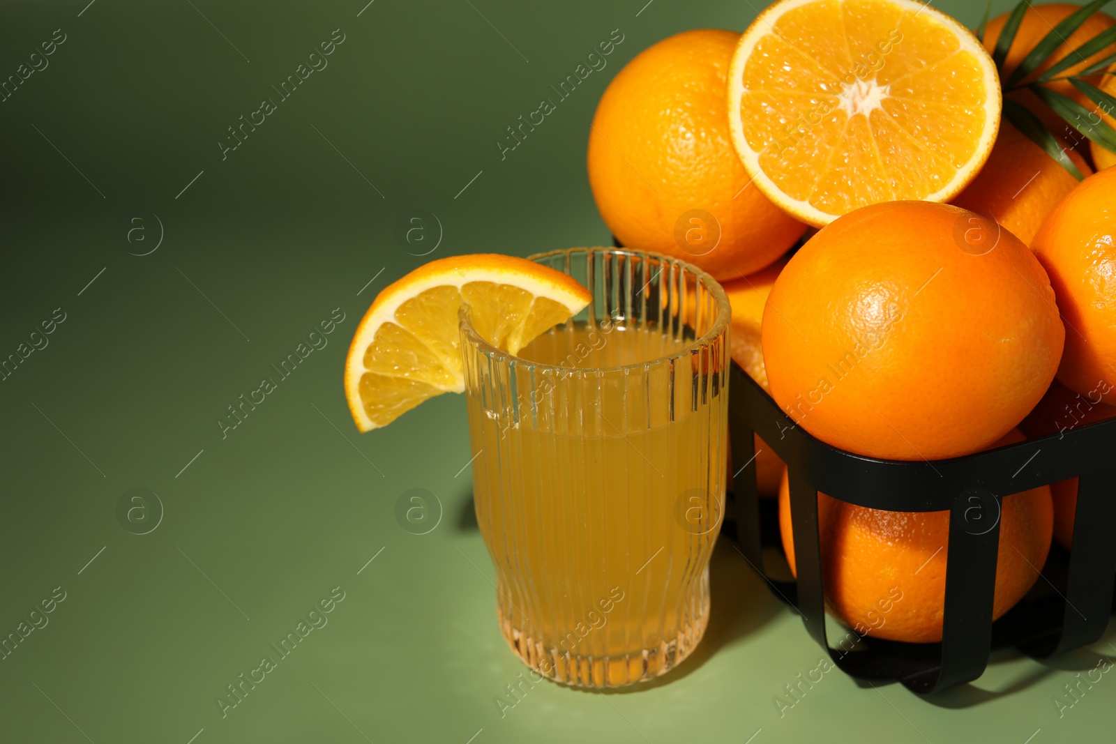 Photo of Fresh oranges in metal basket and glass of juice on green background, closeup. Space for text