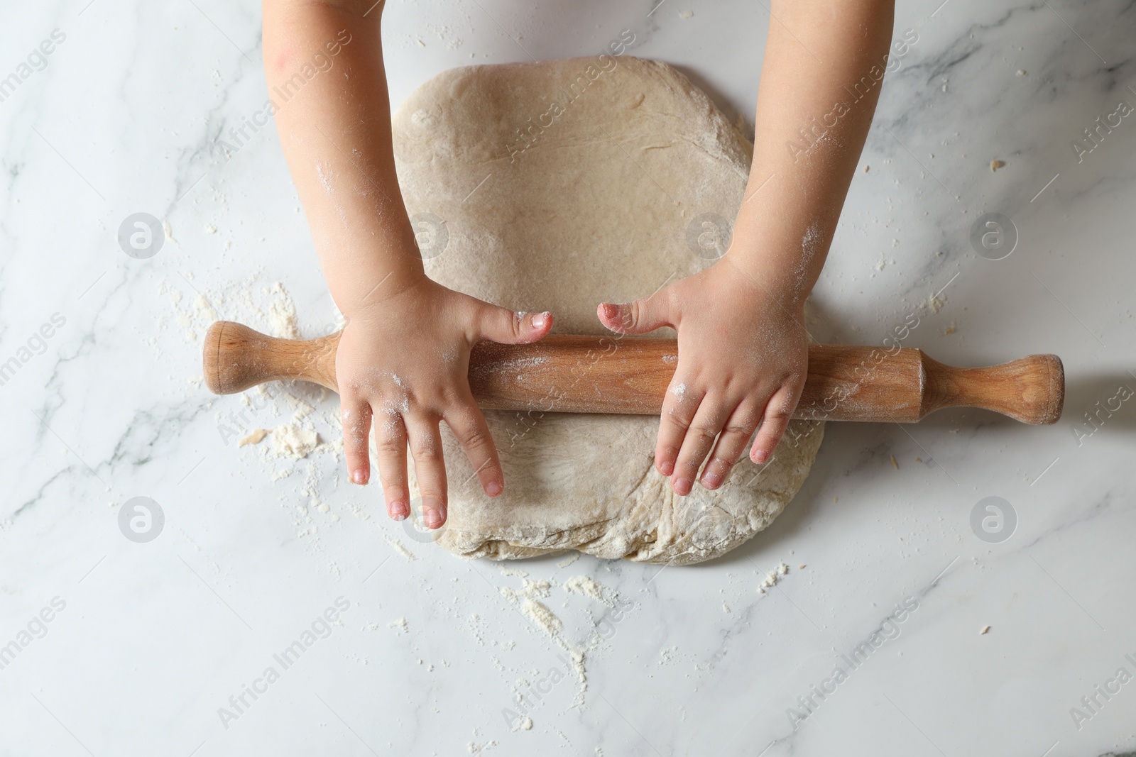 Photo of Little child rolling raw dough at white table, top view