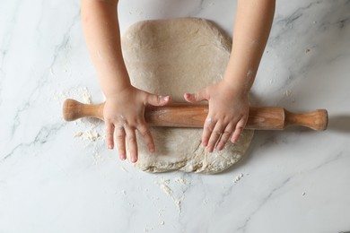 Photo of Little child rolling raw dough at white table, top view