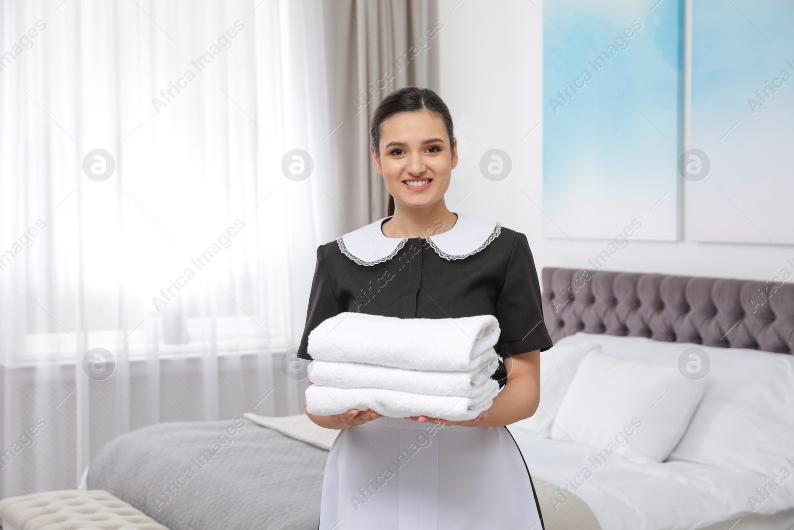 Photo of Young maid holding stack of fresh towels in hotel room