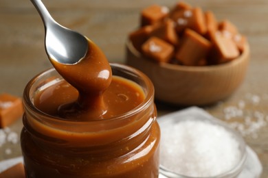 Photo of Taking yummy salted caramel with spoon from glass jar at table, closeup. Space for text