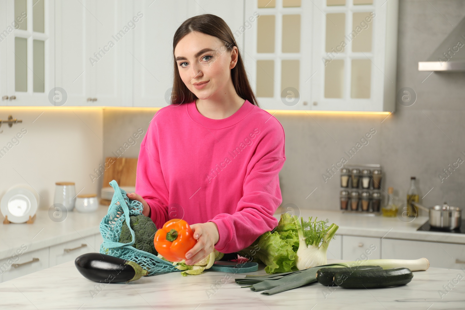 Photo of Woman taking pepper out from string bag at light marble table in kitchen