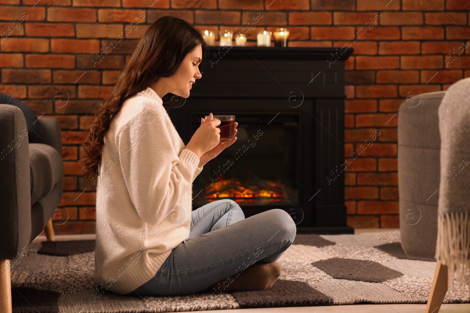 Photo of Young woman with cup of tea relaxing on floor near fireplace at home