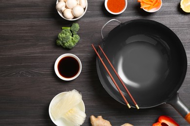 Photo of Empty iron wok, chopsticks and ingredients on dark grey wooden table, flat lay. Space for text