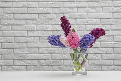 Photo of Beautiful hyacinths in glass vase on table against brick wall, space for text. Spring flowers