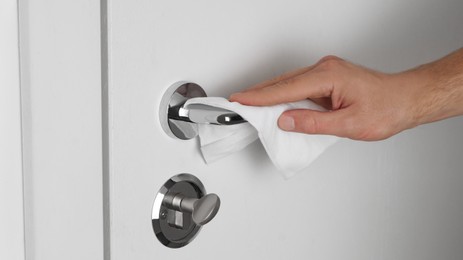 Photo of Man cleaning doorknob with disinfecting wipe indoors, closeup. Protective measures