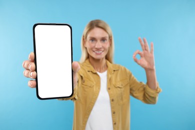 Happy woman holding smartphone with blank screen and showing OK gesture on light blue background, selective focus