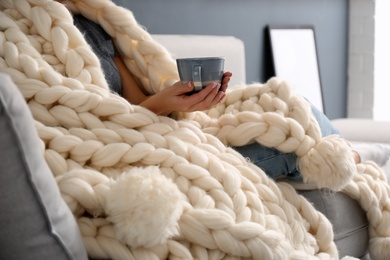 Woman covered with knitted plaid holding cup of coffee on sofa indoors, closeup