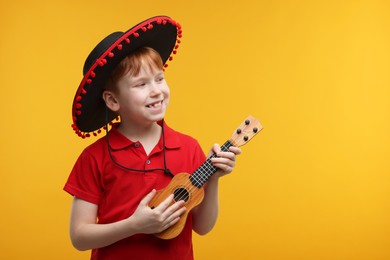 Photo of Cute boy in Mexican sombrero hat playing ukulele on yellow background, space for text