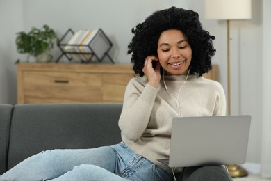 Photo of Happy young woman with earphones using laptop on sofa indoors