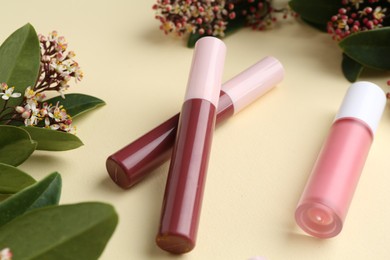 Photo of Different lip glosses, green leaves and flowers on pale yellow background, closeup