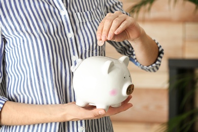 Photo of Woman putting coin into piggy bank indoors, closeup. Money savings