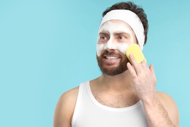 Man with headband washing his face using sponge on light blue background, space for text