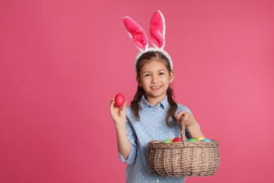Photo of Little girl in bunny ears headband holding basket with Easter eggs on color background, space for text