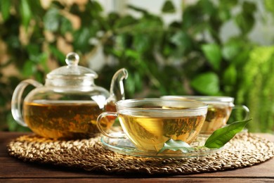 Photo of Fresh green tea in glass cups with saucers, leaves and teapot on wooden table