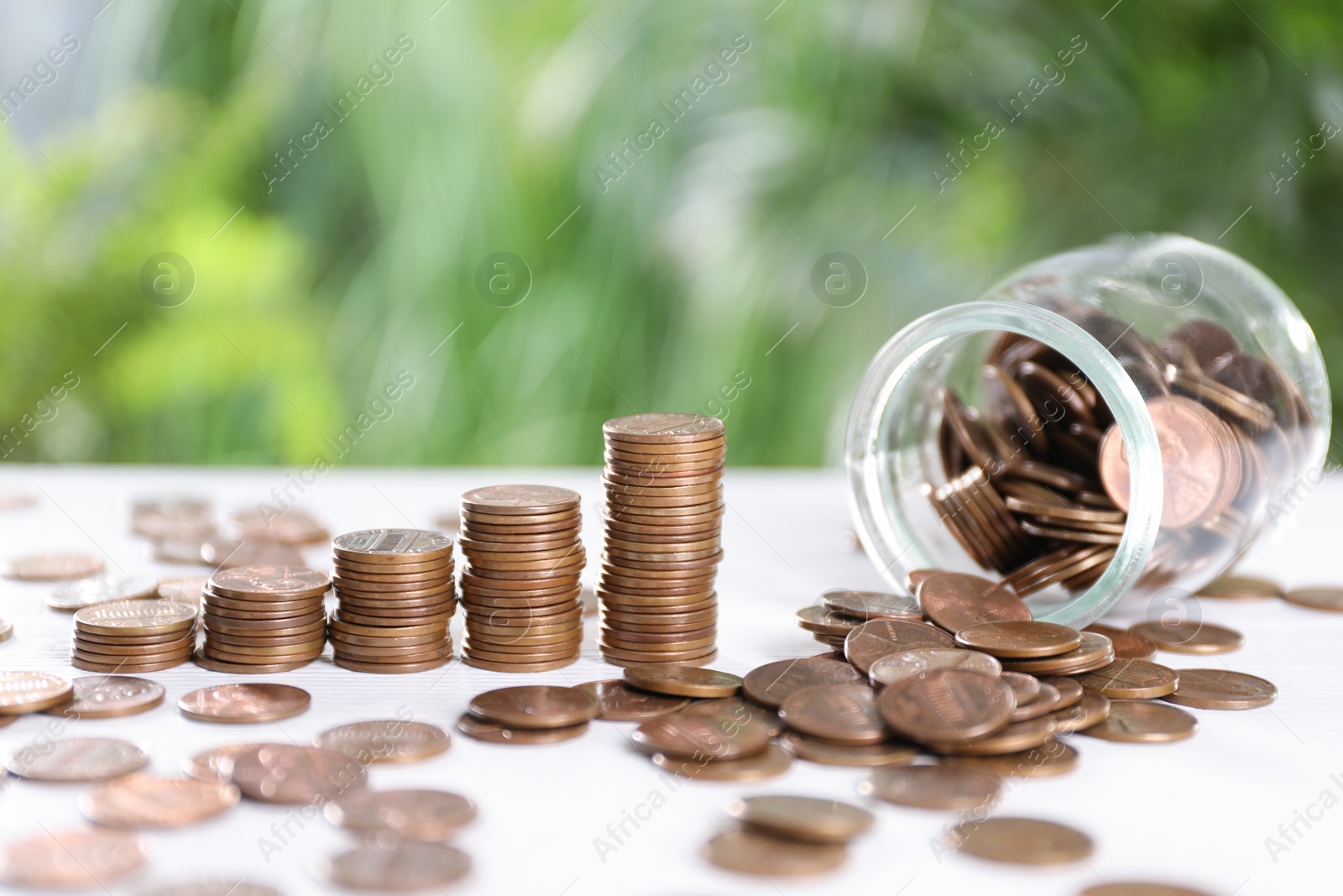 Photo of Overturned glass jar and metal coins on white table against blurred green background. Space for text