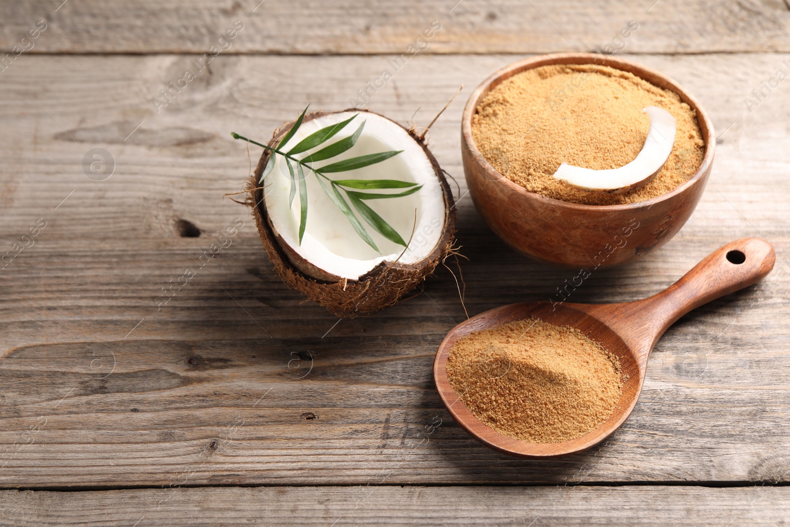 Photo of Coconut sugar in bowl, spoon, fruit and palm leaves on wooden table, closeup. Space for text