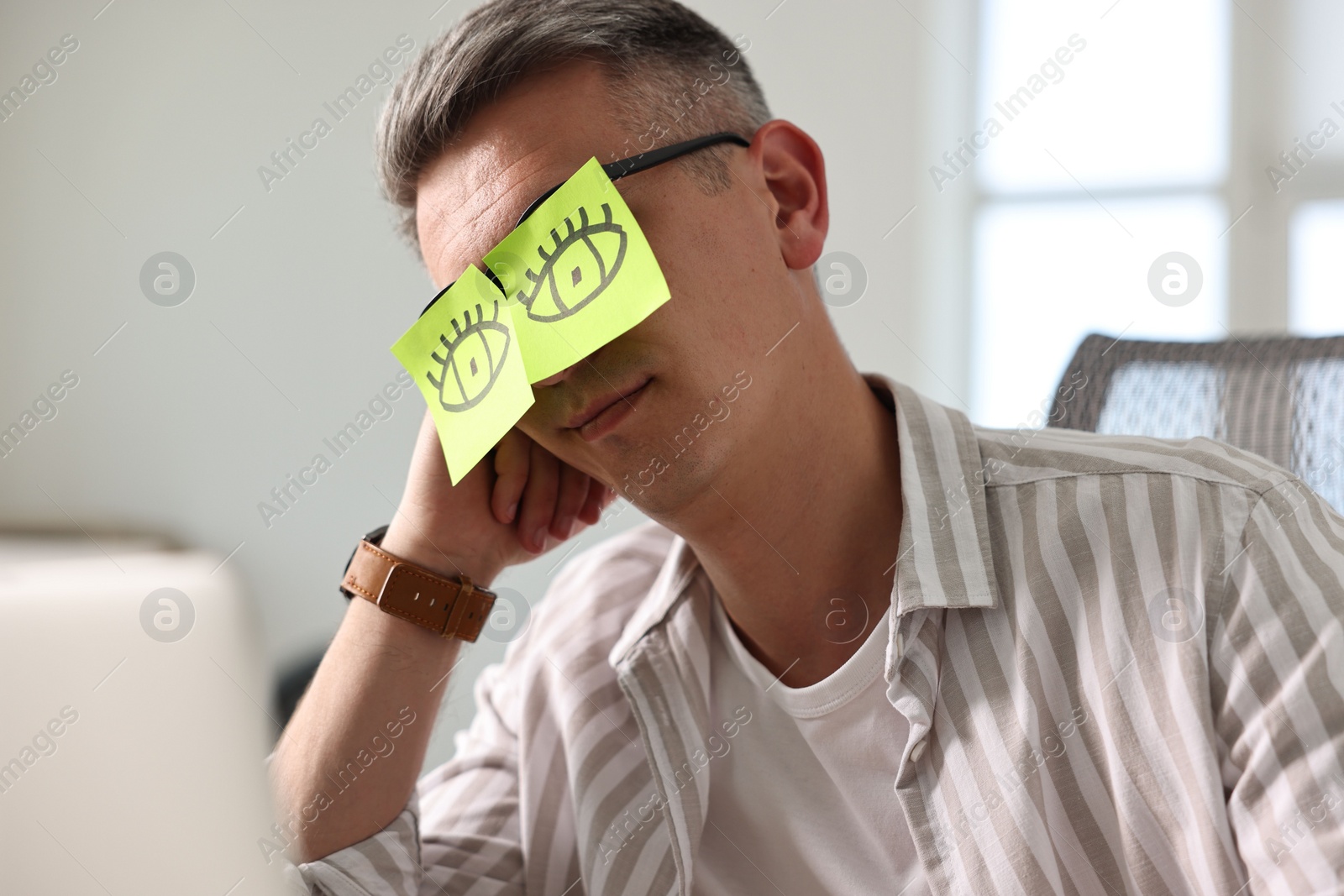 Photo of Man with fake eyes painted on sticky notes snoozing at workplace in office