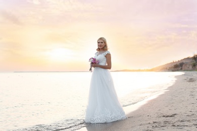 Happy bride holding wedding bouquet on beach at sunset
