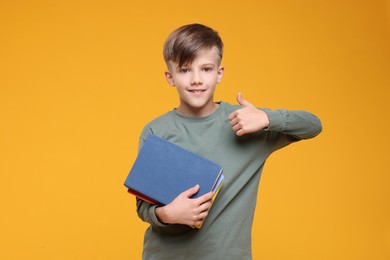 Photo of Cute schoolboy with books showing thumbs up on orange background