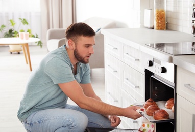Photo of Young man taking out tray of baked buns from oven in kitchen