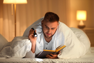 Photo of Young man with flashlight reading book in bedroom