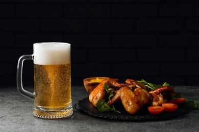 Photo of Mug with beer, delicious baked chicken wings and sauce on grey table