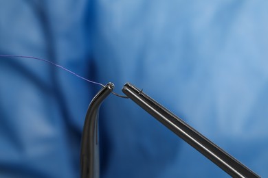 Photo of Forceps with suture thread on blue background, closeup. Medical equipment