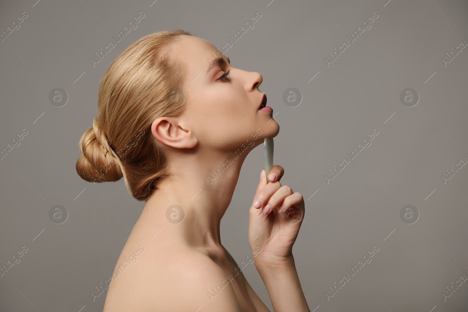 Photo of Beautiful young woman doing facial massage with gua sha tool on grey background