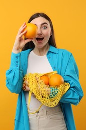 Photo of Woman holding string bag of fresh oranges and covering eye with fruit on orange background