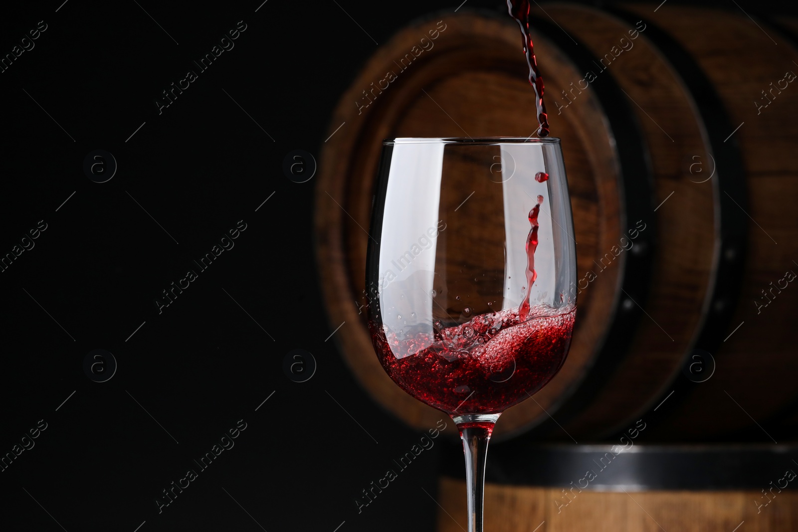 Photo of Pouring red wine into glass near wooden barrels against black background. Space for text