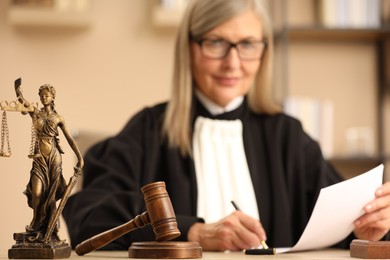 Photo of Judge working indoors, selective focus. Figure of Lady Justice and mallet on wooden table