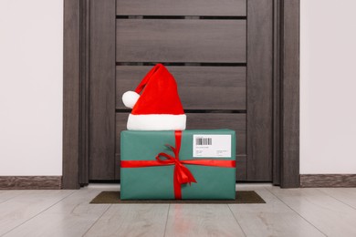 Photo of Christmas gift box and Santa hat near door indoors. Sending present by mail