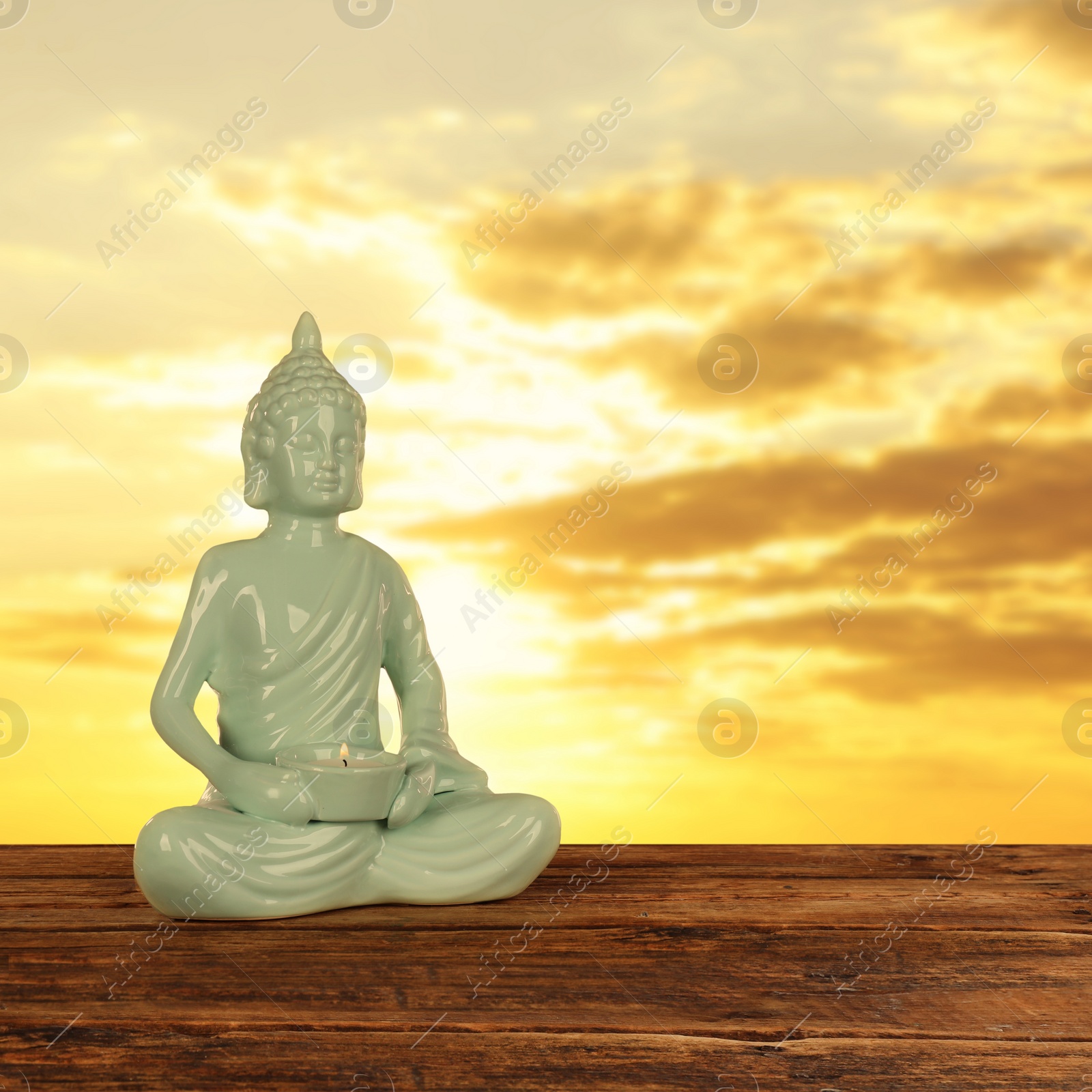 Image of Beautiful ceramic Buddha sculpture with burning candle on wooden surface at sunset. Space for text