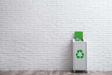 Photo of Overfilled trash bin with recycling symbol near brick wall indoors. Space for text