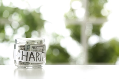 Glass jar with money and word CHARITY on table against blurred background, space for text