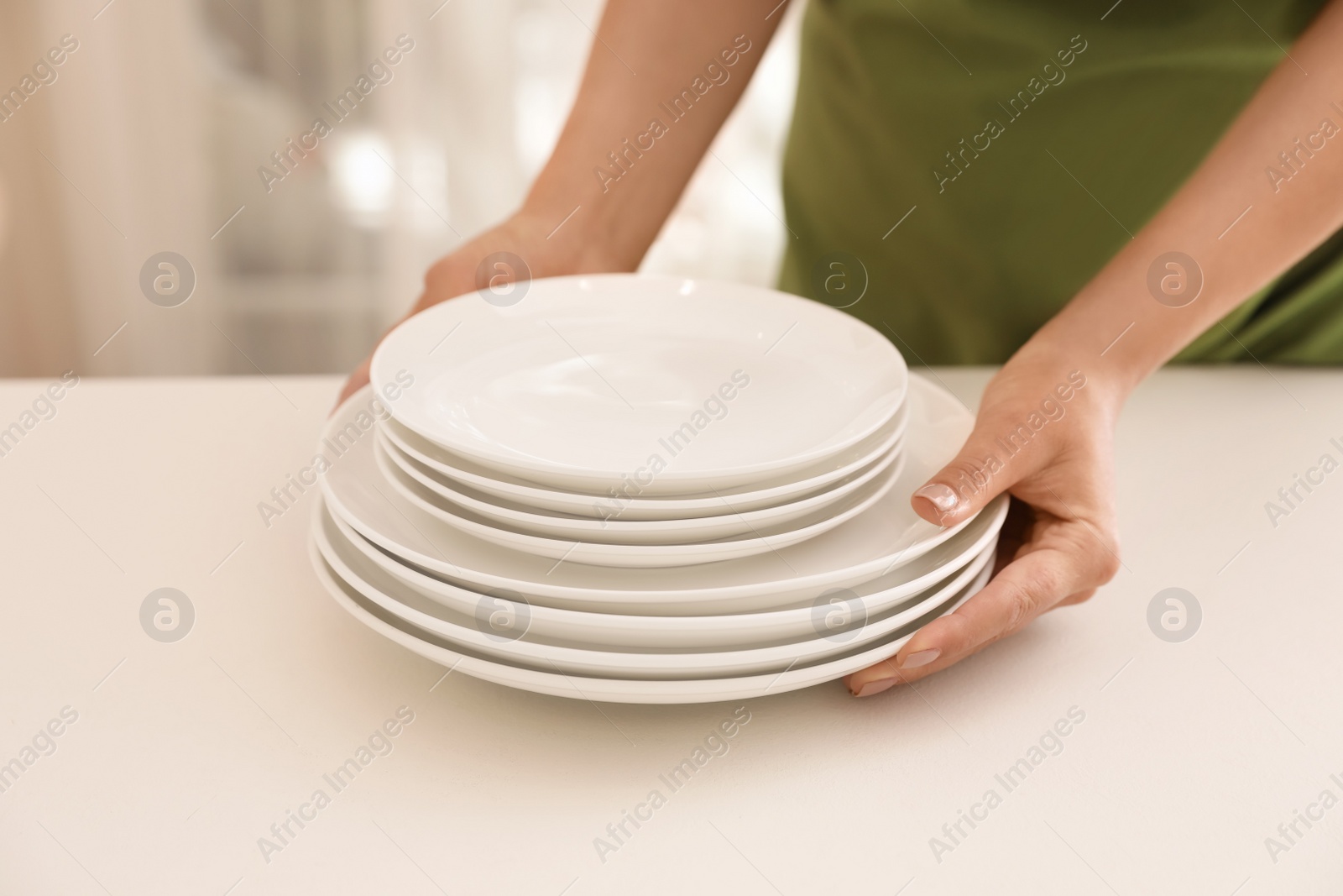 Photo of Woman putting stack of clean dishes on table indoors, closeup