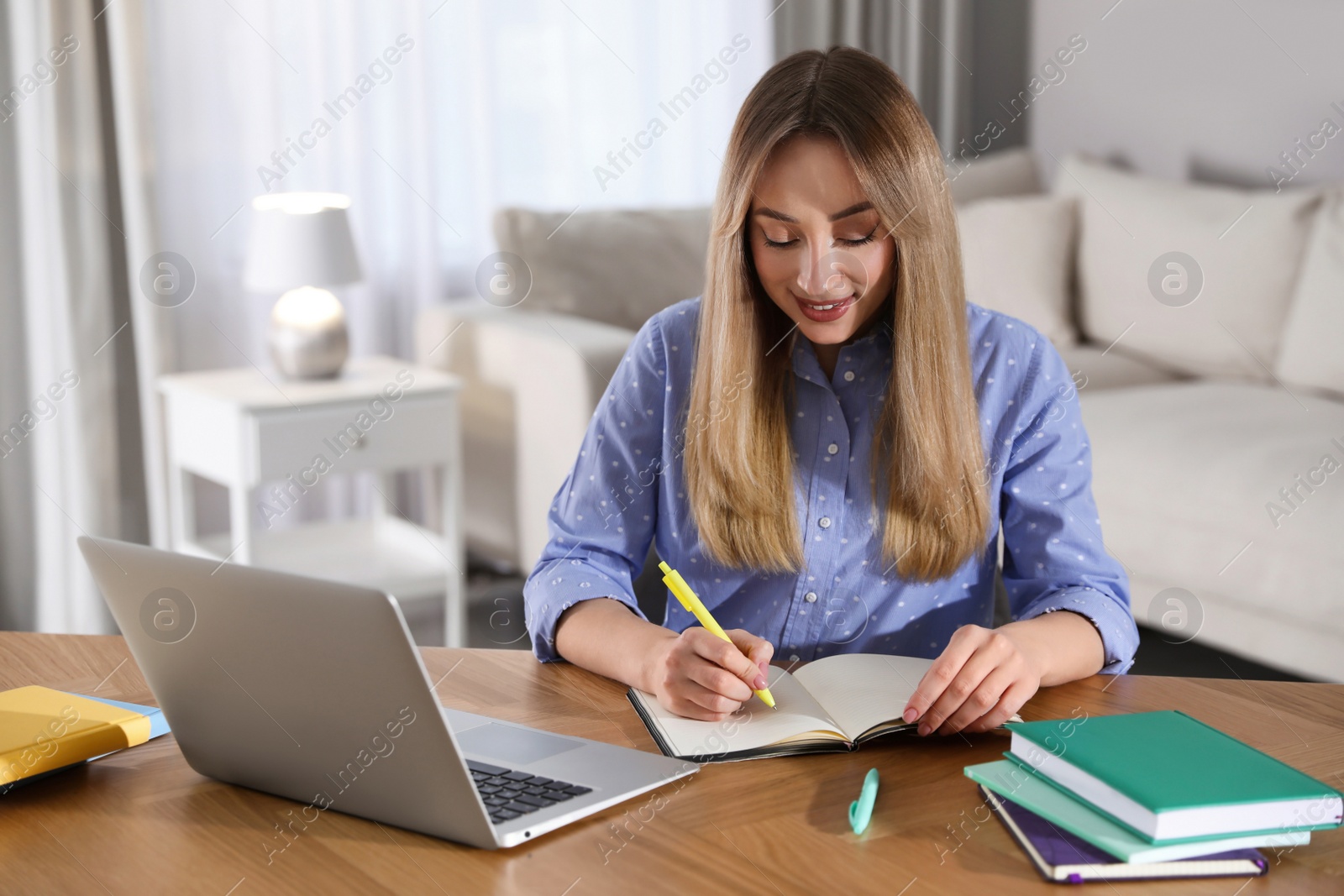 Photo of Young woman writing down notes during webinar at table in room