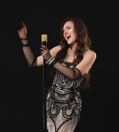 Beautiful young woman in stylish dress with microphone singing on black background, space for text