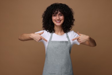 Photo of Happy woman pointing at kitchen apron on brown background. Mockup for design