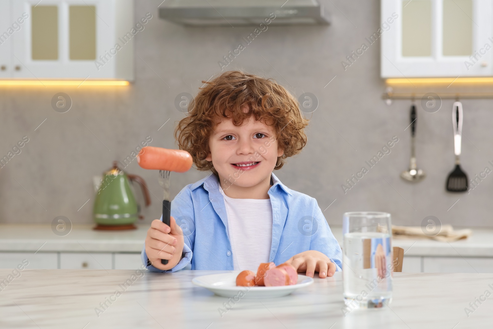 Photo of Cute little boy holding fork with sausages at table in kitchen